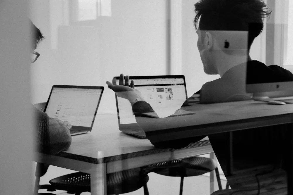 Two men sitting behind glass at a table, staring at laptops and discussing